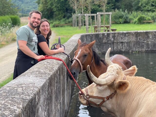 Un couple sourit au bord d’un bassin où Aston boit aux côtés d’un cheval